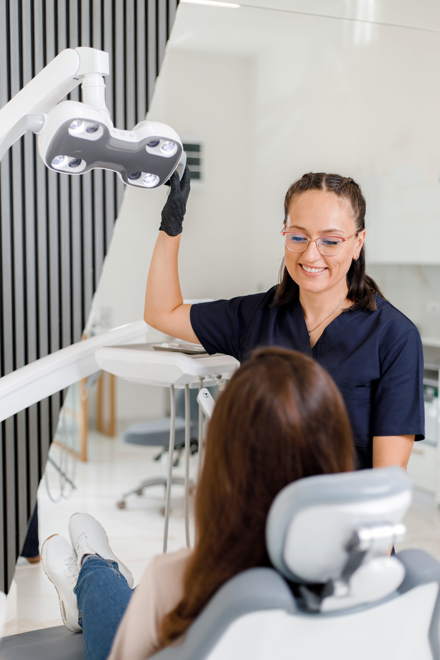 Female dentist in dark scrubs adjusting overhead dental light while smiling at patient in modern clinic treatment room
